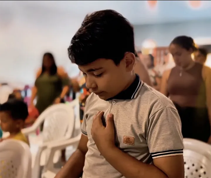 Criança em Adoração - Ministério Infantil El Shaday Parnaíba Menino orando com a mão no peito e olhos fechados durante culto do Ministério Infantil El Shaday Kids em Parnaíba.